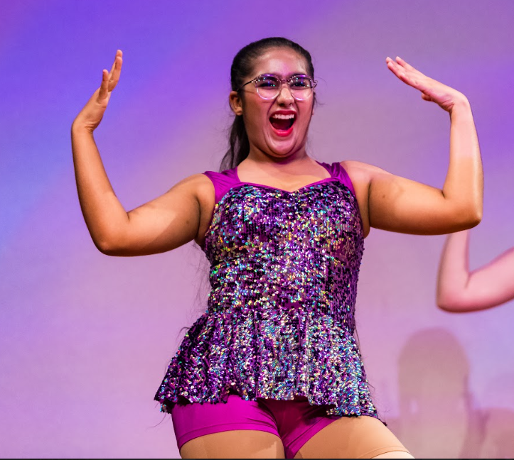 Experienced senior dancer in purple sequin dance costume gracefully posing with hands near ears, radiating joy and confidence during dance class.