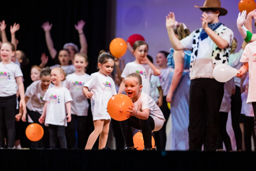 A photo of a happy student from our preschool dance classes catching a orange balloon on stage.